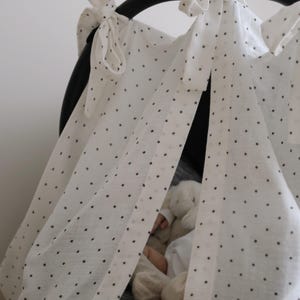 May include: White fabric canopy with black polka dots, tied to a black frame. The canopy is draped over a car seat, with a glimpse of a stuffed animal inside. The fabric is tied with bows.