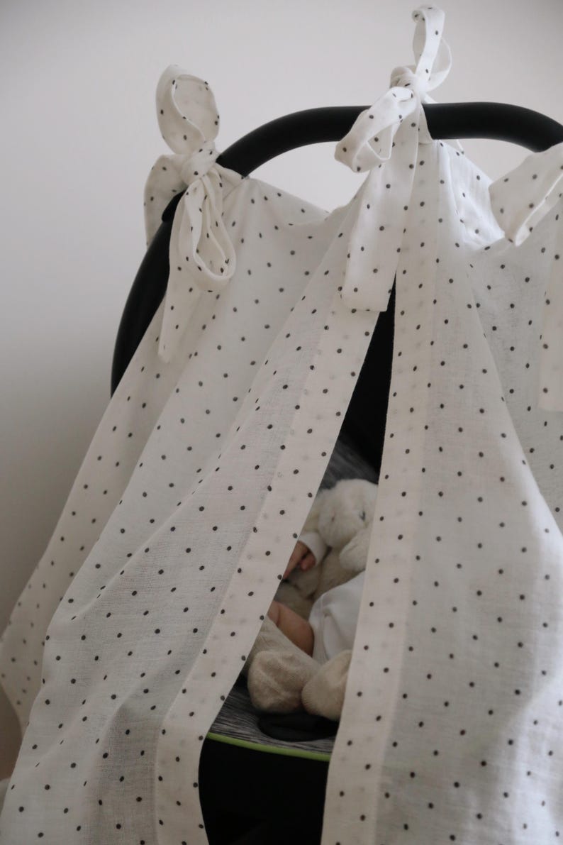 May include: White fabric canopy with black polka dots, tied to a black frame. The canopy is draped over a car seat, with a glimpse of a stuffed animal inside. The fabric is tied with bows.