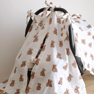 May include: White baby car seat cover with a repeating pattern of brown teddy bears. The cover is tied with bows and drapes over the car seat. A white mug with a wooden spoon is on the wooden surface.