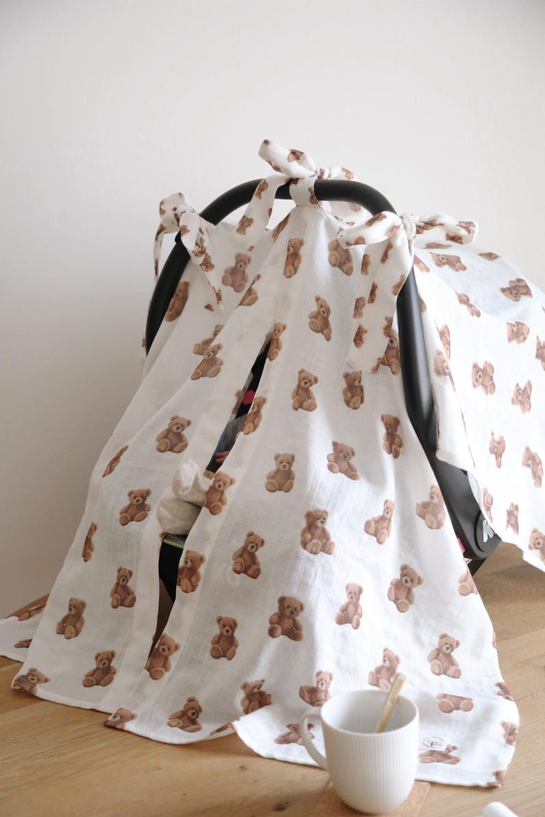 May include: White baby car seat cover with a repeating pattern of brown teddy bears. The cover is tied with bows and drapes over the car seat. A white mug with a wooden spoon is on the wooden surface.