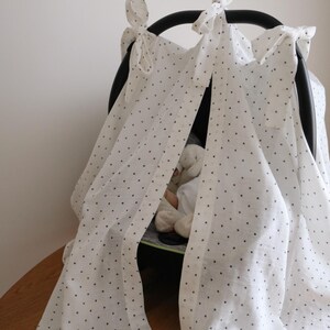 May include: A white baby car seat canopy with black polka dots. The canopy is draped over the car seat frame and tied with bows. A stuffed animal is visible inside the car seat. The canopy is made of a lightweight fabric.