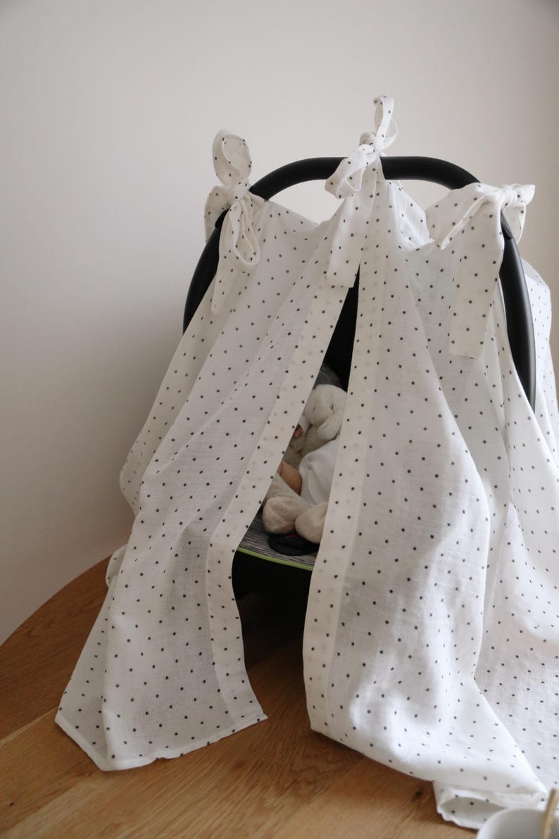 May include: A white baby car seat canopy with black polka dots. The canopy is draped over the car seat frame and tied with bows. A stuffed animal is visible inside the car seat. The canopy is made of a lightweight fabric.