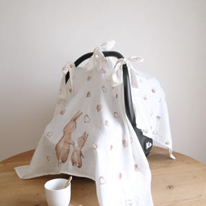May include: White baby car seat cover with a watercolor illustration of two rabbits and scattered brown hearts. The cover is tied to the car seat with white ribbons. A white cup and wooden baby bottle are on the table.