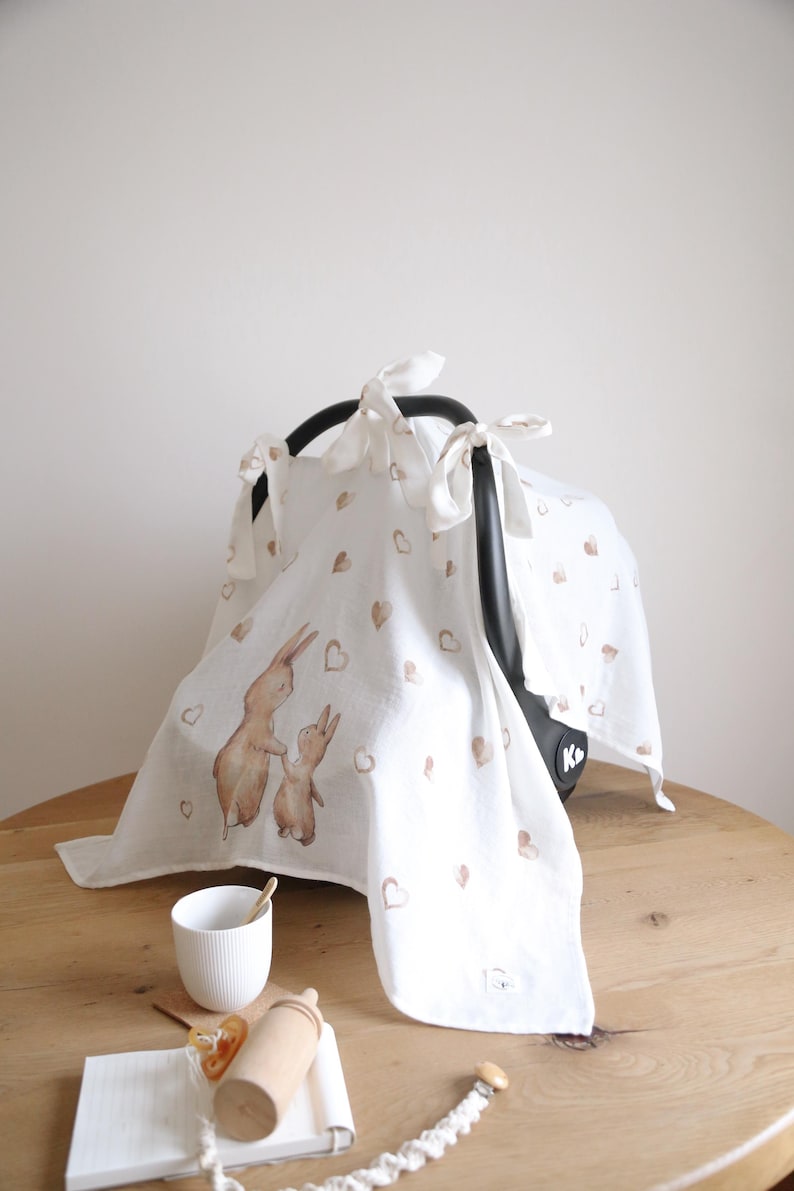 May include: White baby car seat cover with a watercolor illustration of two rabbits and scattered brown hearts. The cover is tied to the car seat with white ribbons. A white cup and wooden baby bottle are on the table.