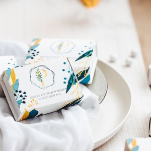 May include: White favor boxes with a blue, yellow, and green floral design. The boxes have the text "Millie & Josiah Pomales 10 October 2021" printed on them.