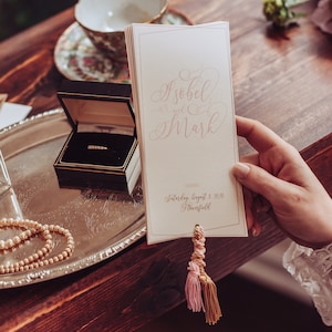 Puede incluir: Una invitación de boda con un borla rosa y dorada. La invitación dice "Isobel y Mark" y "Sábado 1 de agosto de 2020 Thornfield".