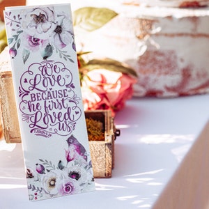 May include: A white bookmark with a floral design and the text "We love because he first loved us 1 John 4:19" in purple lettering.