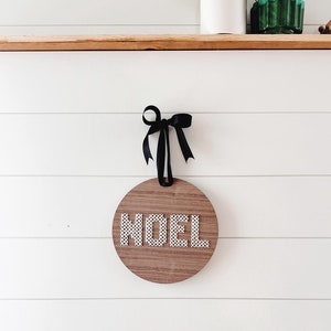 May include: A round wooden sign with the word "NOEL" in white, dotted letters. The sign is suspended by a black ribbon tied in a bow. The background is a white, horizontal-planked wall.
