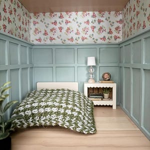 May include: Miniature bedroom scene with a bed covered in a green and white patterned blanket. The walls are painted light blue with floral wallpaper and a small white nightstand with a lamp.