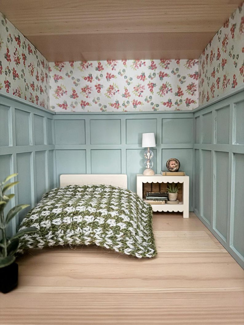 May include: Miniature bedroom scene with a bed covered in a green and white patterned blanket. The walls are painted light blue with floral wallpaper and a small white nightstand with a lamp.