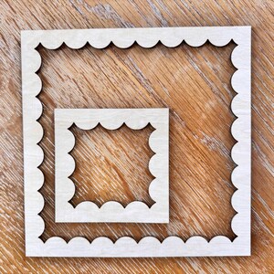 May include: Two unfinished wooden frames with scalloped edges, one nested inside the other. The frames are square and sit on a wooden surface. The wood has a natural, light color and visible grain.