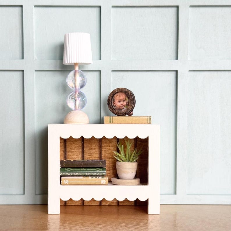May include: A miniature white nightstand with a scalloped edge, holding a lamp with a white shade and iridescent glass orbs. A small framed photo and a stack of books sit on top. The nightstand also has a small potted plant and more books.