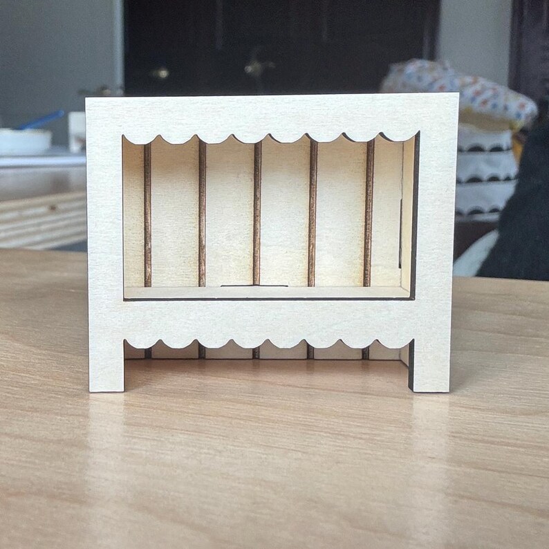 May include: A small, unfinished wooden display shelf with a scalloped top and base. The interior features vertical supports and a small shelf. The natural wood color and simple design suggest a craft or DIY project.