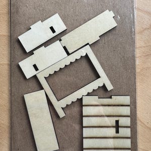 May include: A collection of unfinished wooden pieces for a craft project. The pieces include rectangular and square shapes, a scalloped frame, and parts with slots. The items are arranged on a brown cardboard backing, inside a clear plastic bag.