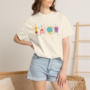 May include: A cream-colored short-sleeve t-shirt featuring a graphic print of four anthropomorphic school supplies: a yellow pencil, red scissors, a blue and green globe, and a purple book, all surrounded by small colorful star illustrations.