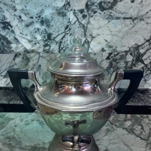 Puede incluir: Una cafetera de plata y vidrio con asas negras. La cafetera tiene un cuerpo redondo con una tapa de vidrio y un pico en la parte delantera.