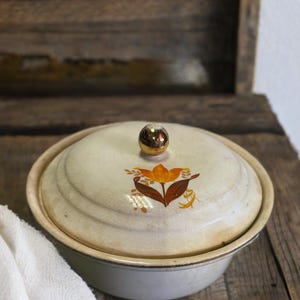 Könnte beinhalten: Eine weiße, abgedeckte Keramikschale mit einem goldenen Knopf auf dem Deckel. Der Deckel hat ein Blumenmuster mit orangefarbenen und braunen Blumen.