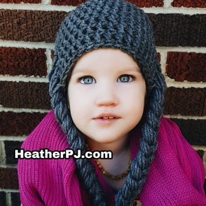 May include: A child wearing a gray knitted hat with long ear flaps and a pink sweater. The hat has a braided design and the child is looking directly at the camera. The text "HeatherPJ.com" is visible in the image.