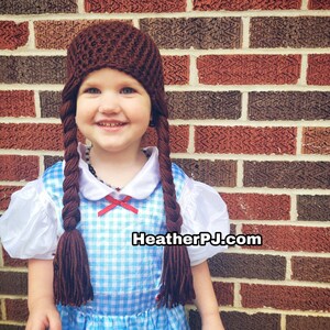 May include: A brown crocheted hat with long braids, worn by a child dressed in a blue and white gingham dress. The hat has a simple design and the braids are long and flowing. The child is smiling and looking at the camera. The background is a brick wall. HeatherPJ.com is visible in the image.