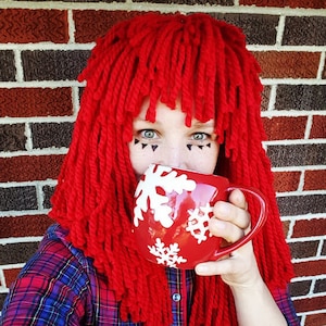 May include: A red mug with white snowflake designs is held up to the face. The person is wearing a red yarn wig and a plaid shirt. The background is a brick wall.