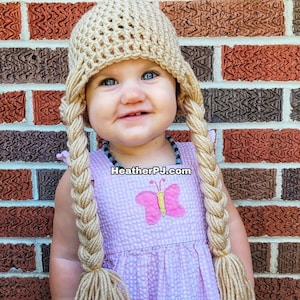May include: A young child wearing a beige crocheted hat with long braids. The hat has a butterfly design on the front. The child is wearing a pink and white dress with a butterfly design. The child is standing in front of a brick wall.
