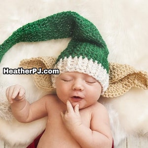 May include: A newborn baby wearing a green and white elf hat with brown ears. The hat has a long green tail.