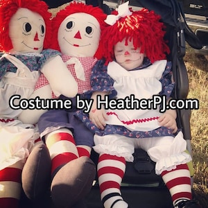 May include: A baby dressed as a Raggedy Ann doll, sitting in a stroller with two large Raggedy Ann dolls. The baby is wearing a red and white striped dress, a white apron, and a red wig. The dolls are wearing red and white striped dresses and have red hair.