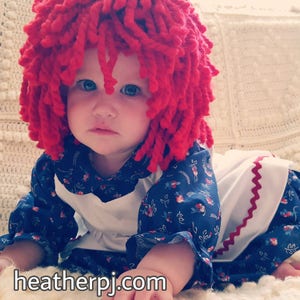 May include: A baby wearing a red yarn wig and a blue and white floral dress with a white apron and red trim. The baby is crawling on a white crocheted blanket.