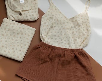 May include: A set of clothing featuring a cream-colored button-down shirt and matching folded fabric, both with a small floral pattern. Also included is a cream-colored camisole with the same floral print and solid brown elastic-waist shorts.