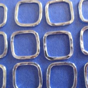 May include: A close-up of several silver-toned metal square rings with a hammered texture. The rings are arranged in rows on a blue surface.