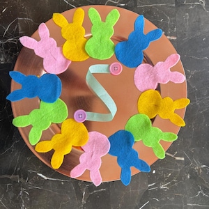May include: A copper-colored decorative plate adorned with colorful felt bunny cutouts in pink, yellow, green, and blue. A light blue ribbon and two pink buttons add detail to the Easter-themed craft.