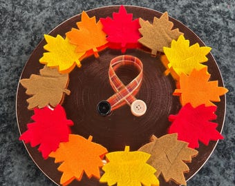 May include: A circular brown plate decorated with felt maple leaves in shades of red, orange, yellow, and brown. In the center, a plaid ribbon is looped into a circle and secured with one black button and one beige button, creating a decorative accent.