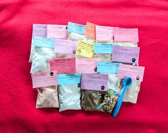 May include: A collection of 10 small bags of cake mix and frosting with instructions on each bag. The bags are on a red surface with a blue spoon.