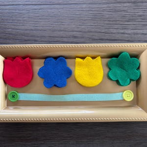 May include: A beige rectangular tray with a beaded border holds four colorful felt shapes: a red tulip, a blue flower, a yellow tulip, and a green flower. A light blue ribbon with green and yellow buttons is below the shapes.