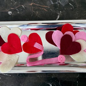 May include: A silver tray filled with felt hearts in shades of red, pink, and cream. A pink and white checkered ribbon and pink buttons add detail. The arrangement is a decorative display.