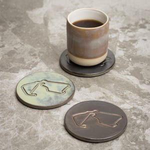 May include: A ceramic mug with coffee rests on a gray coaster. Two additional coasters with a racetrack design are visible. One coaster is light green, the other is gray. The coasters are round and have a recessed design.