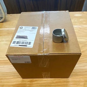May include: A brown cardboard shipping box with a USPS Ground Advantage shipping label and a small silver metal tin with a matching lid placed on top. The label includes a barcode and text, and a secondary label reads 2 oz. Candle Tin.