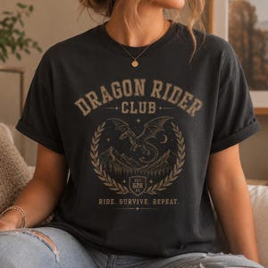 Può includere: Una t-shirt grigio antracite con una stampa grafica che riporta il testo "DRAGON RIDER CLUB" e "RIDE. SURVIVE. REPEAT.". Il design include un drago in volo, una catena montuosa e una corona d'alloro con uno scudo "EST. 628 AD.".