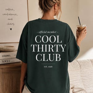May include: A dark green short-sleeve t-shirt featuring white text on the back that reads: - official member - COOL THIRTY CLUB est. 1996. The shirt has a crew neckline and a relaxed, loose-fitting silhouette with a plain front and back design.
