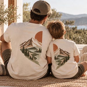 Könnte beinhalten: Zwei cremefarbene T-Shirts mit einem Grafikdruck auf der Rückseite. Das Design zeigt ein Gebirge, einen gewundenen Fluss, immergrüne Bäume und eine große orangefarbene Sonne. Die T-Shirts werden von zwei Personen getragen, eine trägt eine beigefarbene Kappe.