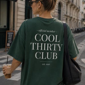 May include: A dark green short-sleeve t-shirt featuring white text on the back that reads - official member - COOL THIRTY CLUB est. 1996. The shirt has a loose, relaxed fit with a crew neckline and is worn with light blue denim jeans.