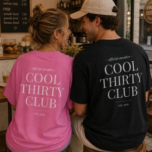 May include: A bright pink t-shirt and a black t-shirt, both featuring the white text "official member COOL THIRTY CLUB est. 1996" printed on the back. The shirts have a classic crew neck design and short sleeves, displayed from a rear-facing perspective.