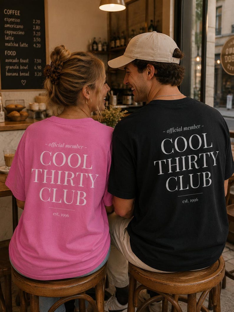 May include: A bright pink t-shirt and a black t-shirt, both featuring the white text "official member COOL THIRTY CLUB est. 1996" printed on the back. The shirts have a classic crew neck design and short sleeves, displayed from a rear-facing perspective.