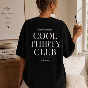 May include: A black short-sleeved t-shirt featuring white text on the back that reads - official member - COOL THIRTY CLUB est. 1996. The shirt has a relaxed fit and is worn with light-colored pants.