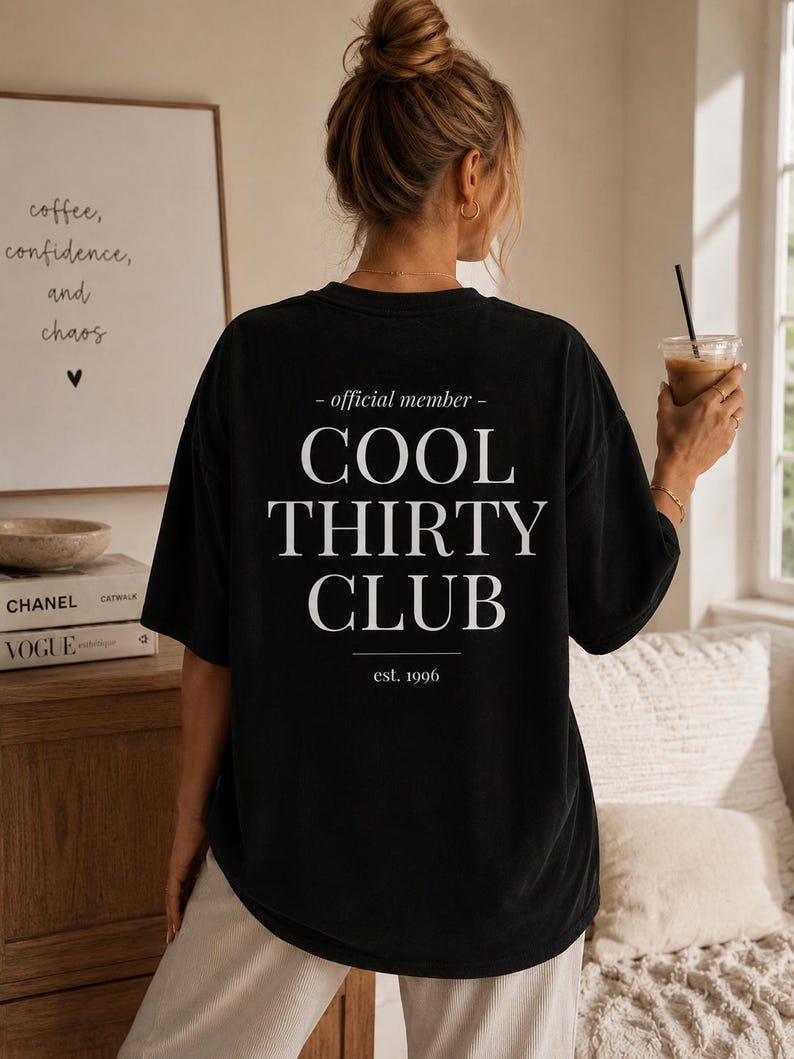 May include: A black short-sleeved t-shirt featuring white text on the back that reads - official member - COOL THIRTY CLUB est. 1996. The shirt has a relaxed fit and is worn with light-colored pants.