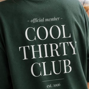 May include: A dark forest green t-shirt featuring white text on the back that reads - official member - COOL THIRTY CLUB est. 1996. The design is centered with a clean serif font, including horizontal lines above and below the main text.
