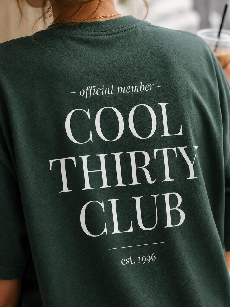 May include: A dark forest green t-shirt featuring white text on the back that reads - official member - COOL THIRTY CLUB est. 1996. The design is centered with a clean serif font, including horizontal lines above and below the main text.
