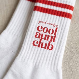 Könnte beinhalten: Weiße Socken mit roten Streifen am oberen Rand. Der Text "proud member cool aunt club" ist rot aufgedruckt. Die Socken bestehen aus geripptem Strickmaterial.