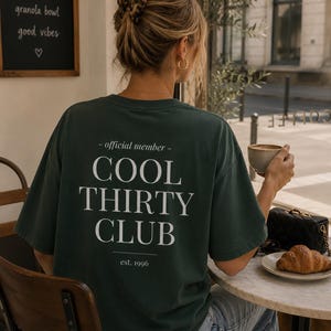 May include: A dark green short-sleeved t-shirt featuring white text on the back that reads - official member - COOL THIRTY CLUB est. 1996. The shirt has a crew neckline and a relaxed fit, worn with light blue denim jeans.