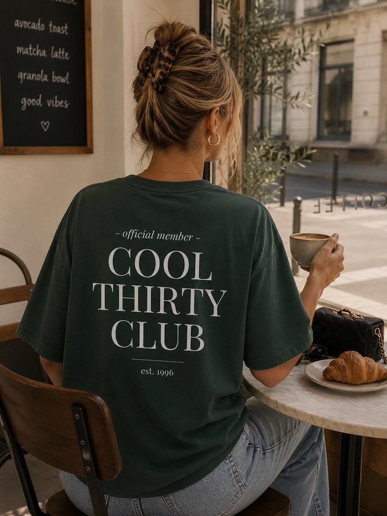 May include: A dark green short-sleeved t-shirt featuring white text on the back that reads - official member - COOL THIRTY CLUB est. 1996. The shirt has a crew neckline and a relaxed fit, worn with light blue denim jeans.